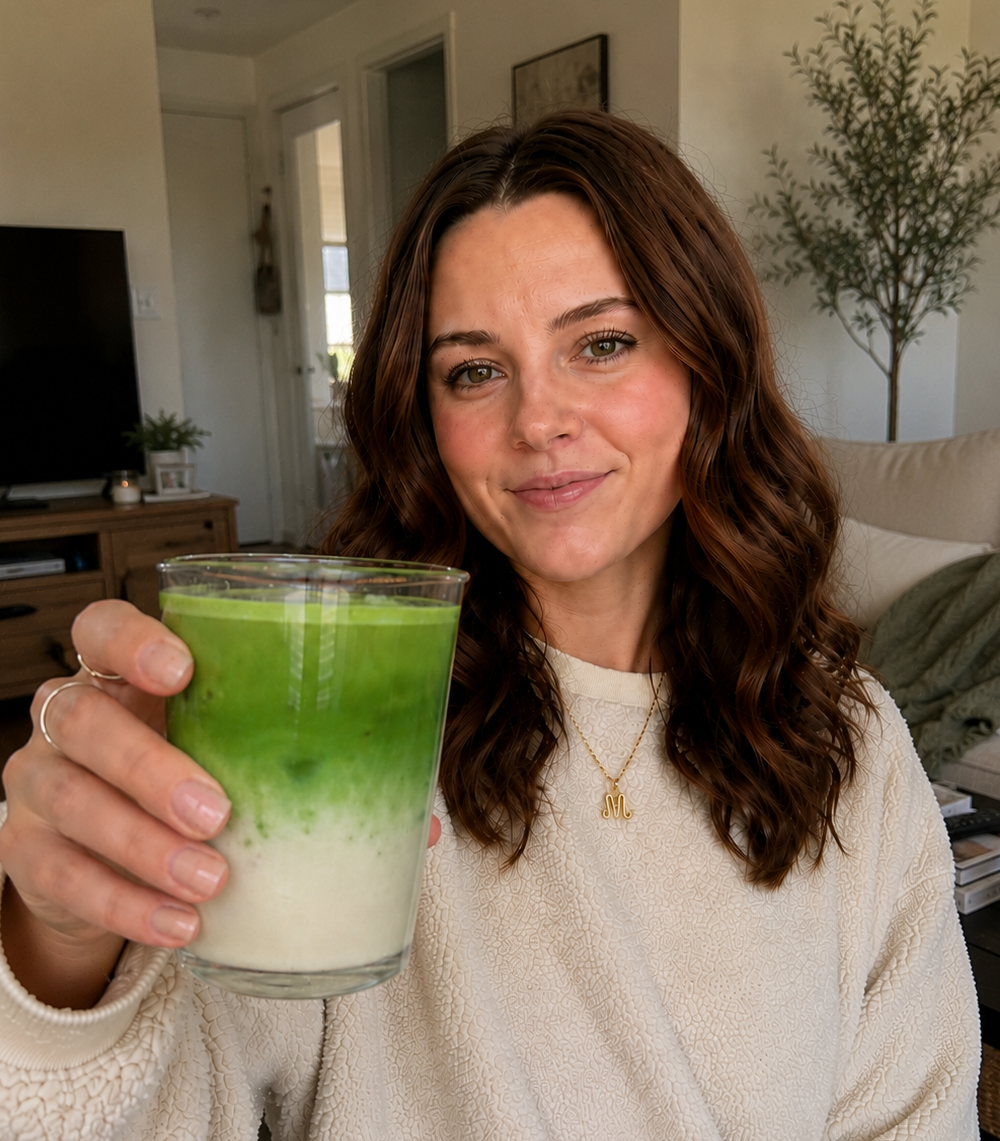 A woman with wavy brown hair holds up a glass of layered green and white beverage in a cozy indoor setting. Ceremonial grade matcha reviews 