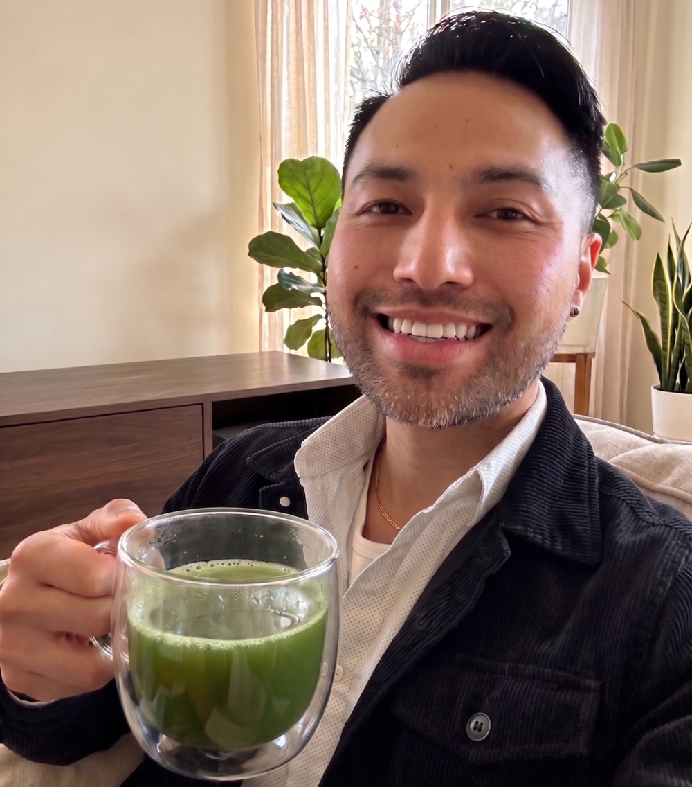 A smiling man holding a clear double-walled glass mug filled with japanese ceremonial grade matcha, wearing a black corduroy jacket over a white shirt in a brightly lit indoor setting.