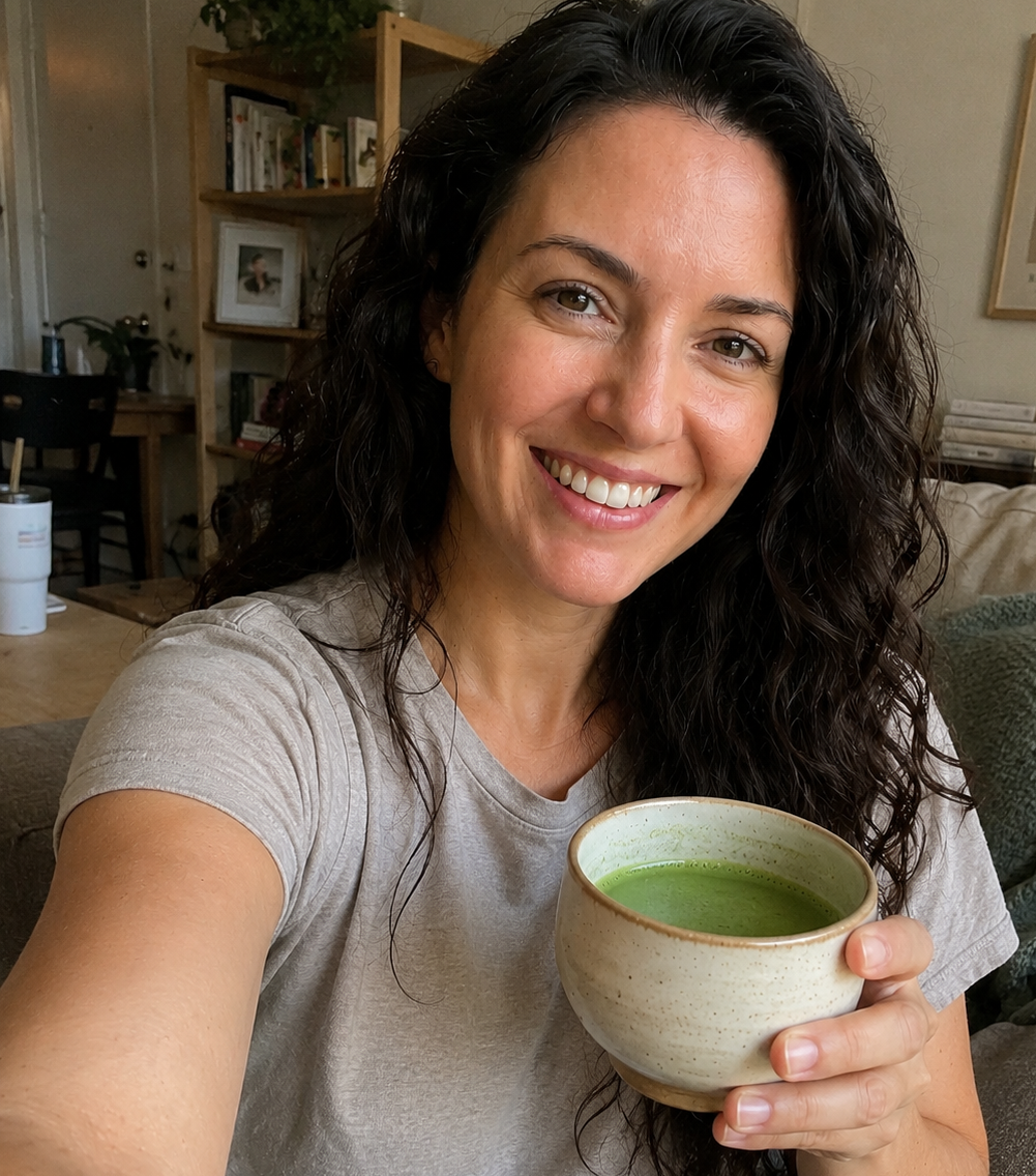 A smiling woman with long dark curly hair takes a selfie while holding a handmade ceramic cup filled with japanese ceremonial grade matcha in a living room